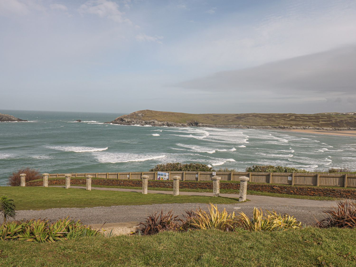 A coastal view with waves crashing and a grassy area with plants and a stone fence at Guillemot in Crantock