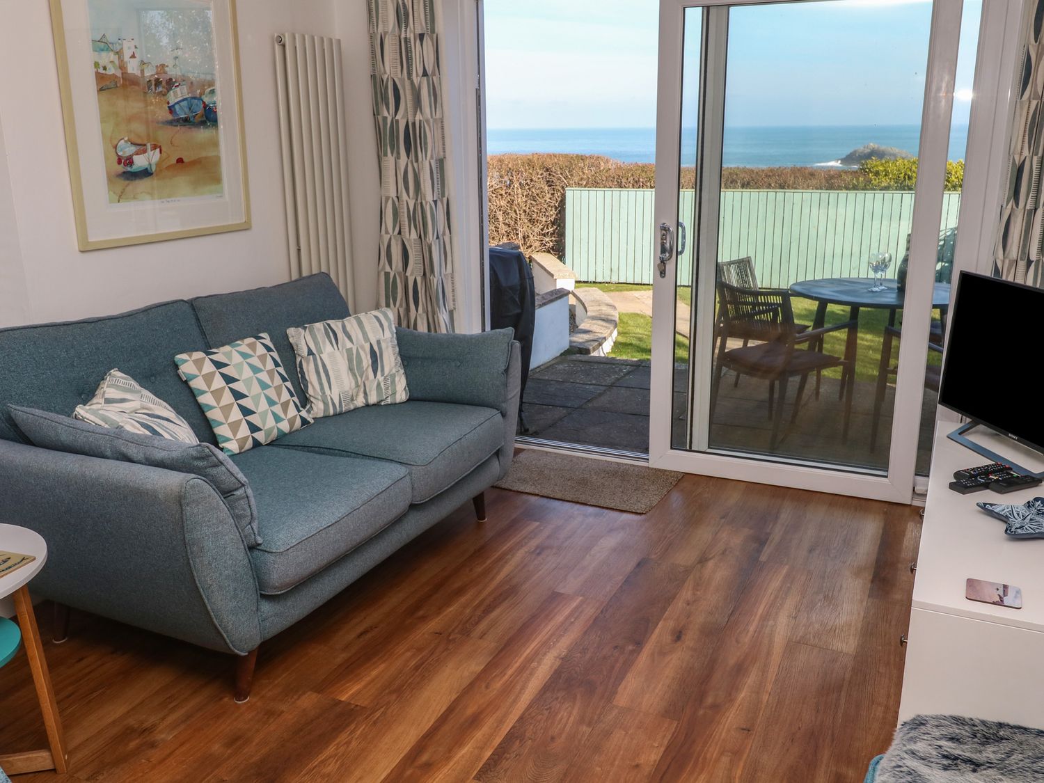A living room with a blue sofa and geometric patterned cushions next to sliding glass doors opening to a garden and sea view at Kittiwake in Crantock