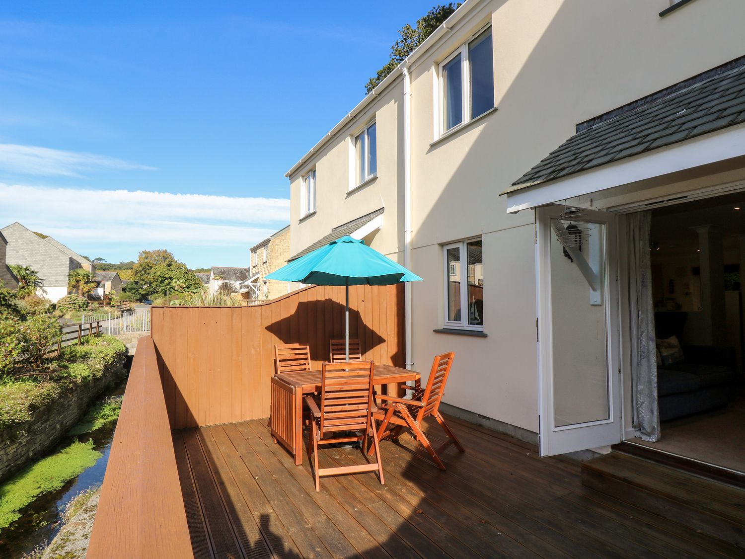 A wooden outdoor deck with a table and four chairs under a blue umbrella next to a cream colored house at Fox's Walk in Falmouth