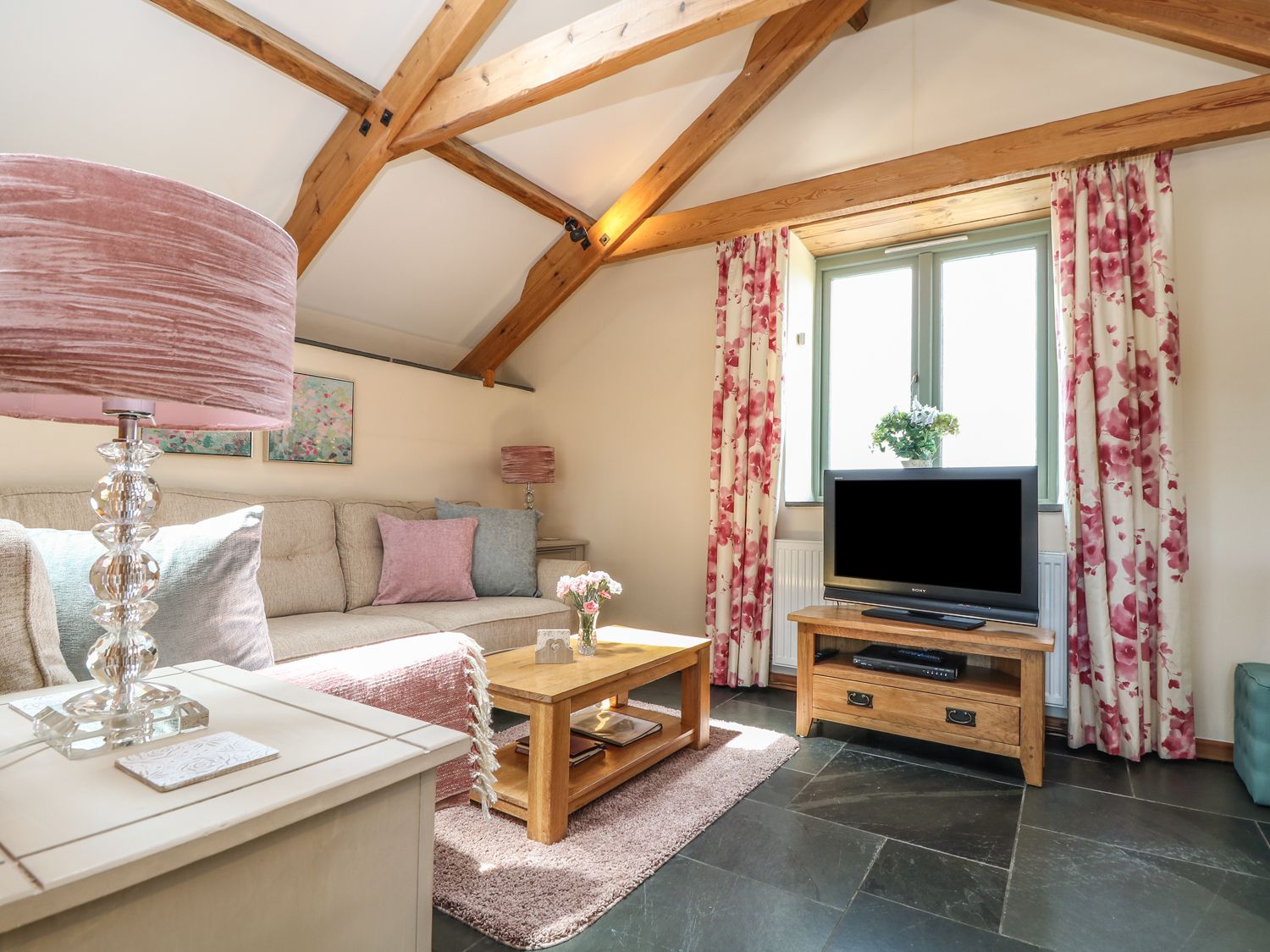 A living room with a beige sofa pink and blue cushions wooden coffee table tv on wooden stand and floral curtains at Oak Cottage in Tregony near Mevagissey