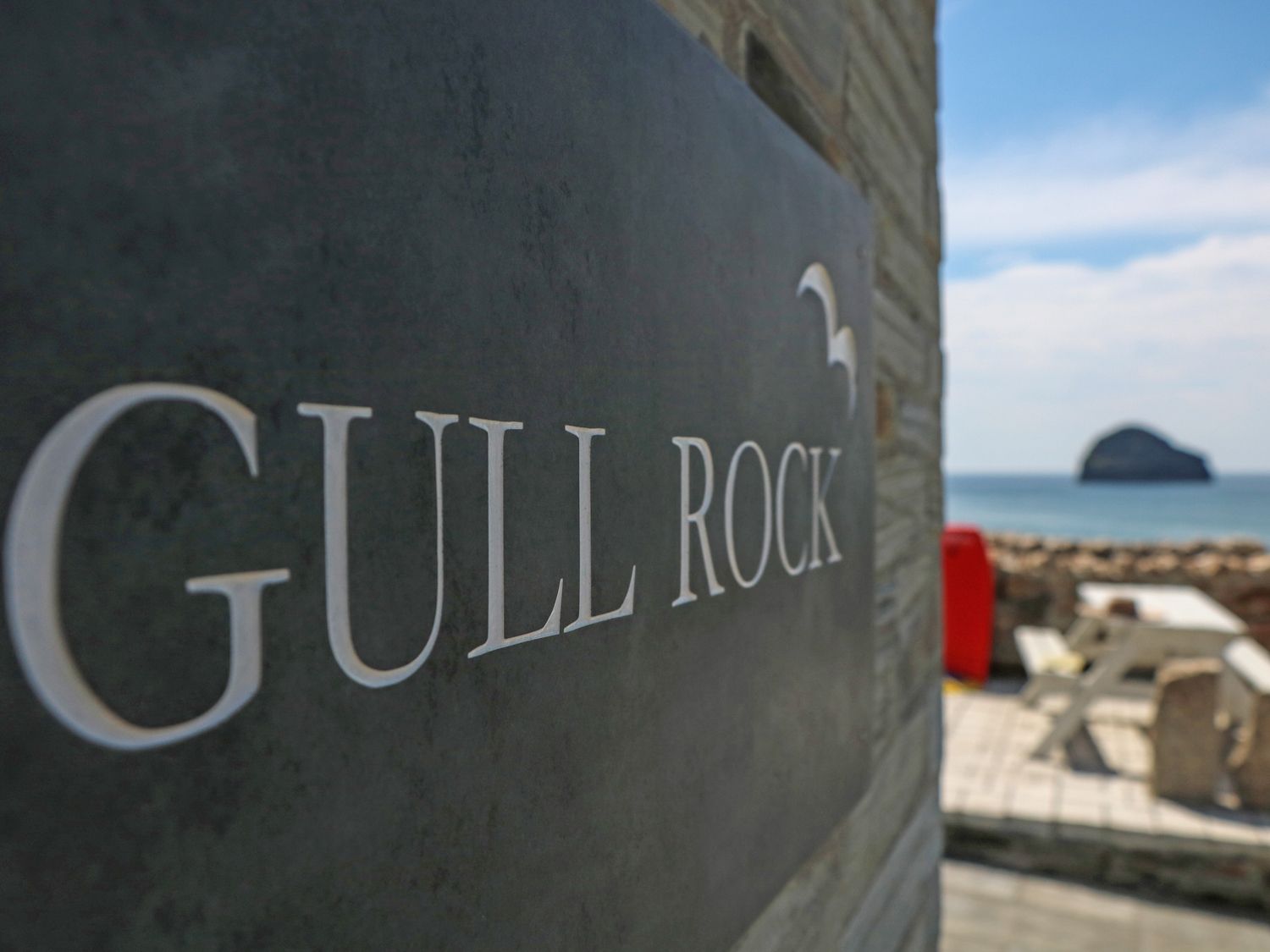 A sign with Gull Rock text on a wall near an outdoor seating area and ocean view at Gull Rock in Trebarwith Strand