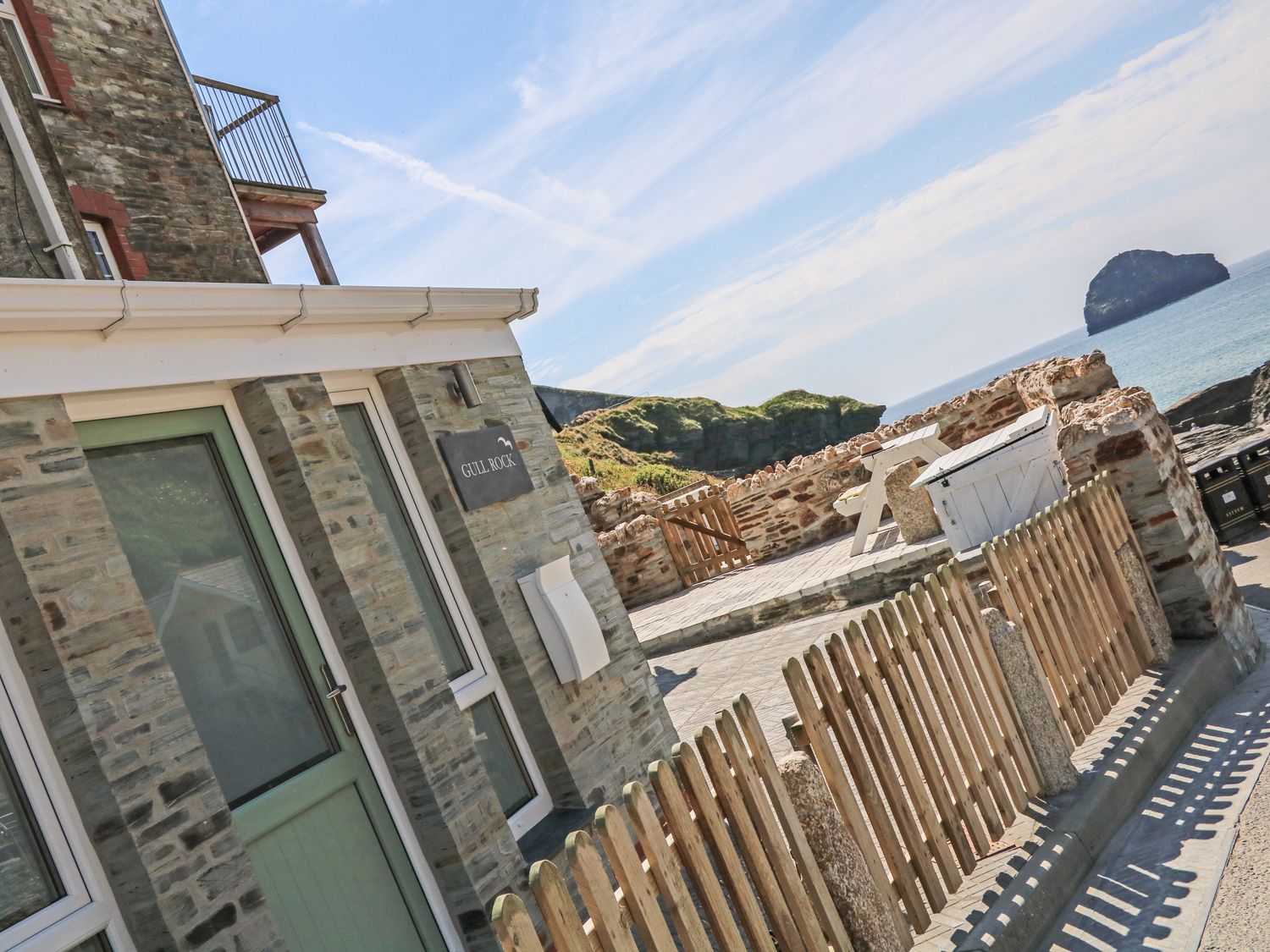A stone building with a green door and windows next to a wooden fence and a stone wall with a picnic table overlooking the sea at Gull Rock Trebarwith Strand