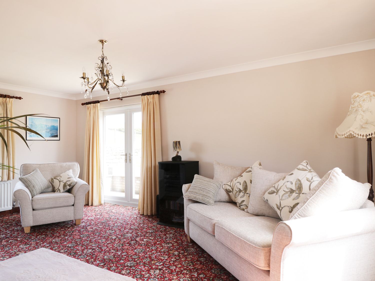 A living room with a sofa, armchair, red patterned carpet, chandelier, and curtains at Acorns in Brampton