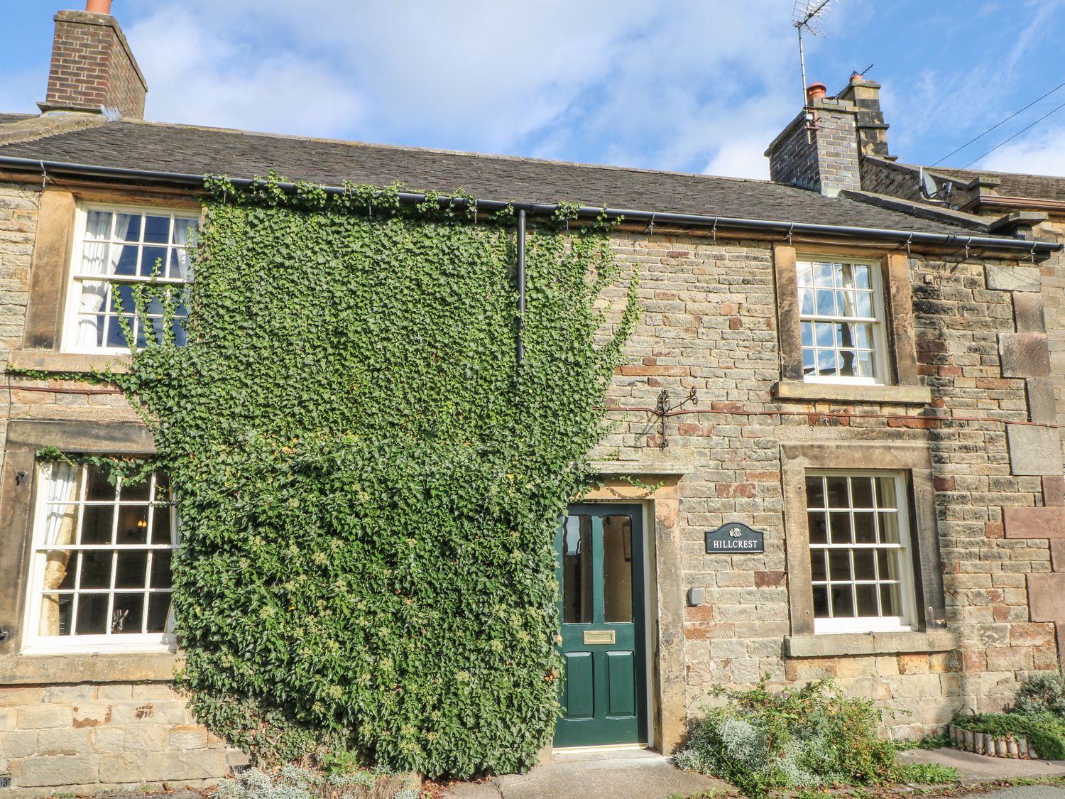 The front of a stone house with green ivy growing over part of the wall and a green door at Hillcrest in Longnor