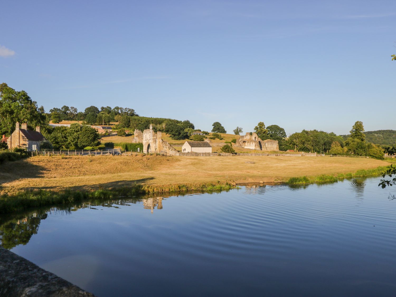 A river in front of dry grass fields with old stone ruins and houses in the background at Oak Lodge in Sheriff Hutton