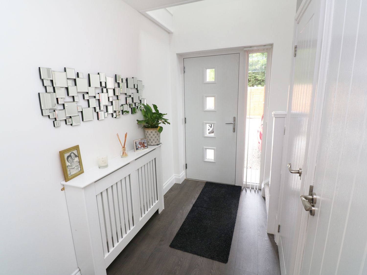 A hallway with a grey front door a black rug a white radiator cover with framed photos a plant and a decorative mirror on the wall at Little Pippin in Carlyon Bay near St Austell