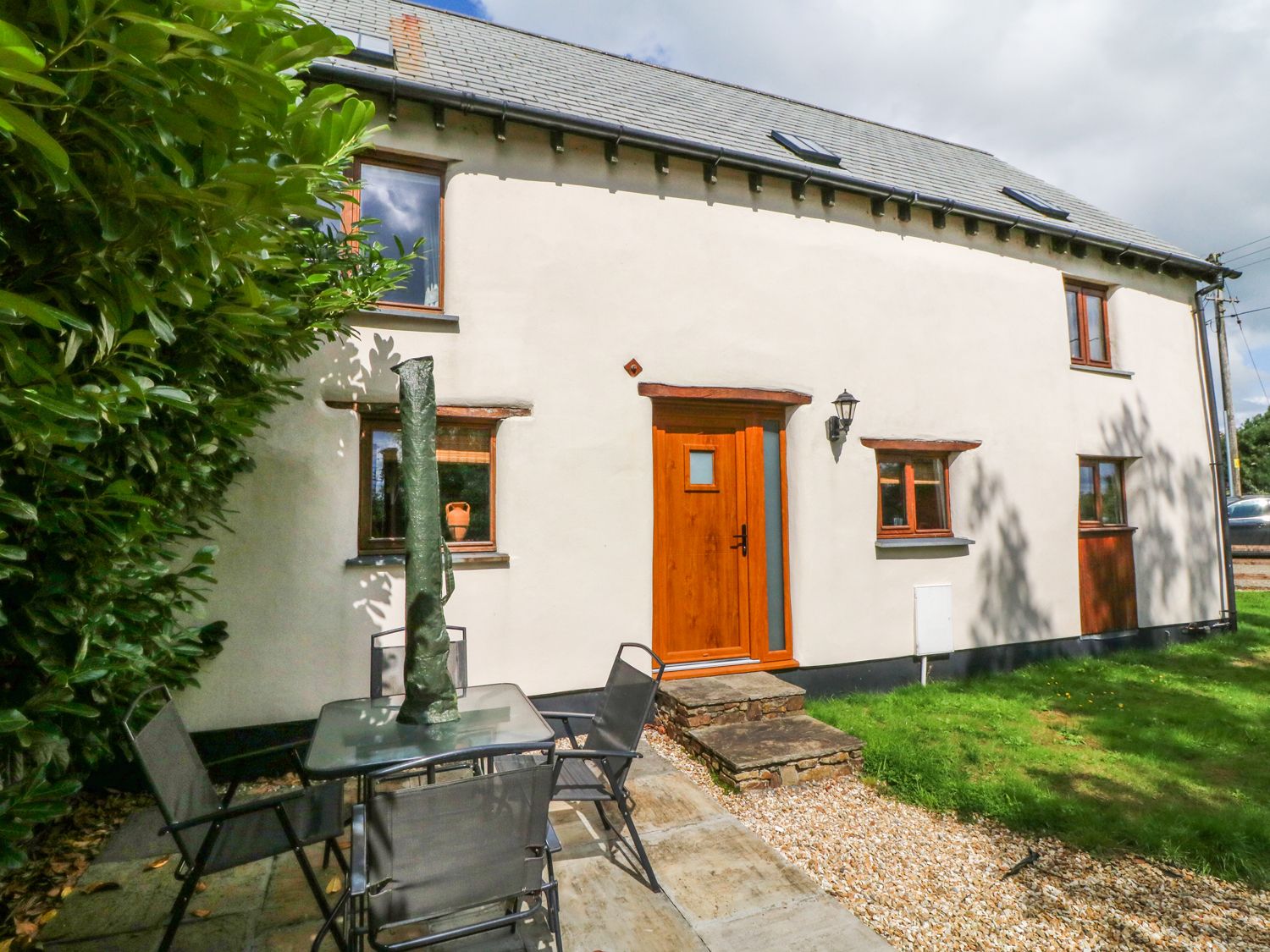An outdoor patio with a glass table and four chairs next to a house with a wooden door and several windows at Post House in Holsworthy