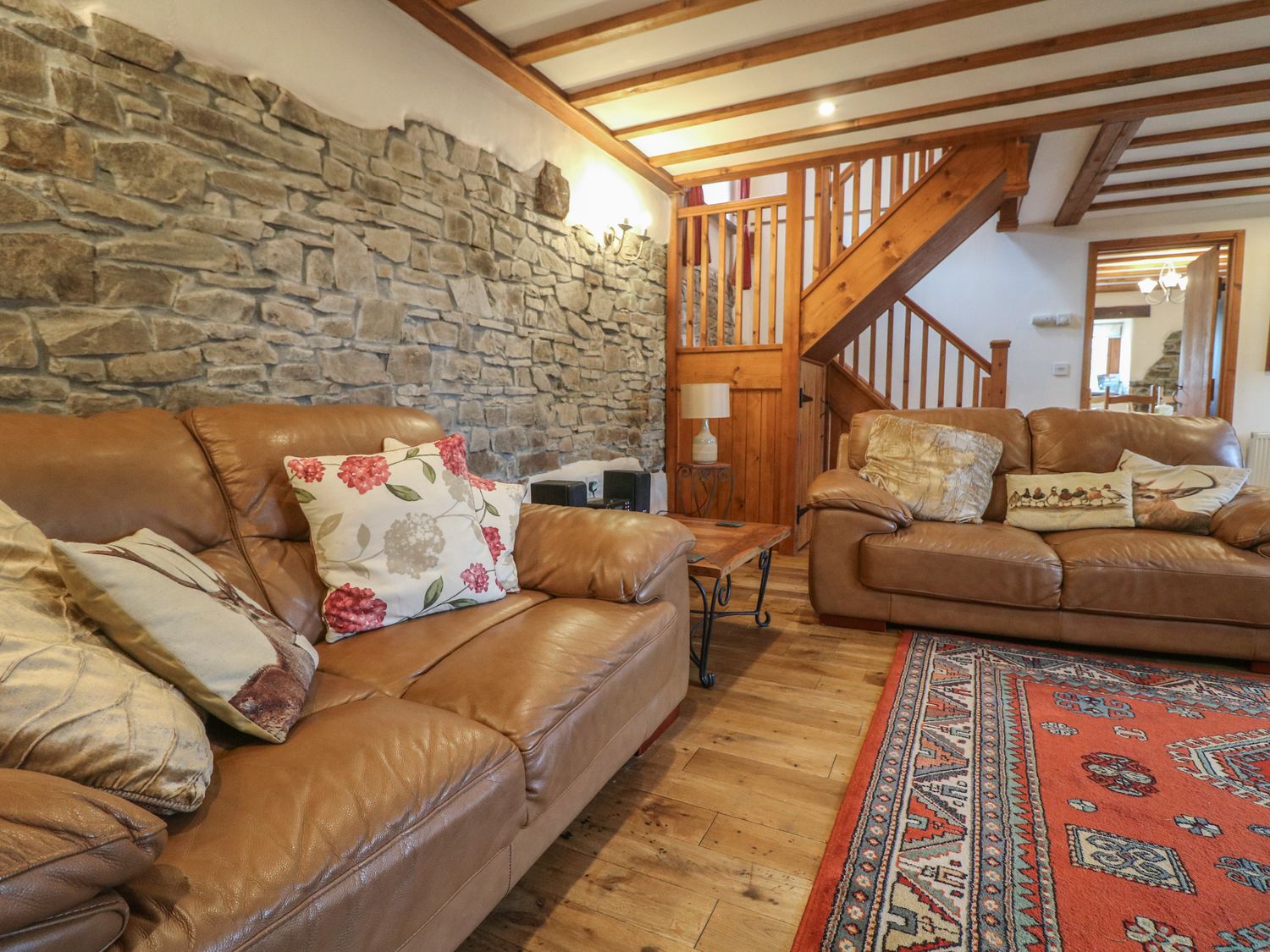 A living room with brown leather sofas and patterned cushions next to a stone wall and wooden staircase at Post House in Holsworthy