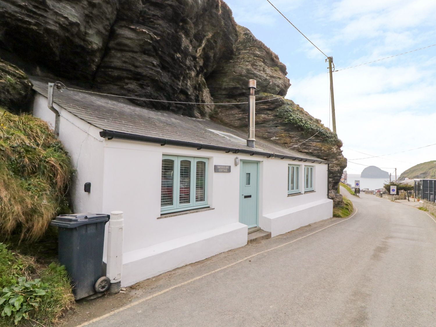 A white cottage built against a rock face with a pale blue door and windows on a narrow road at Smugglers Cottage Trebarwith Strand near Tintagel