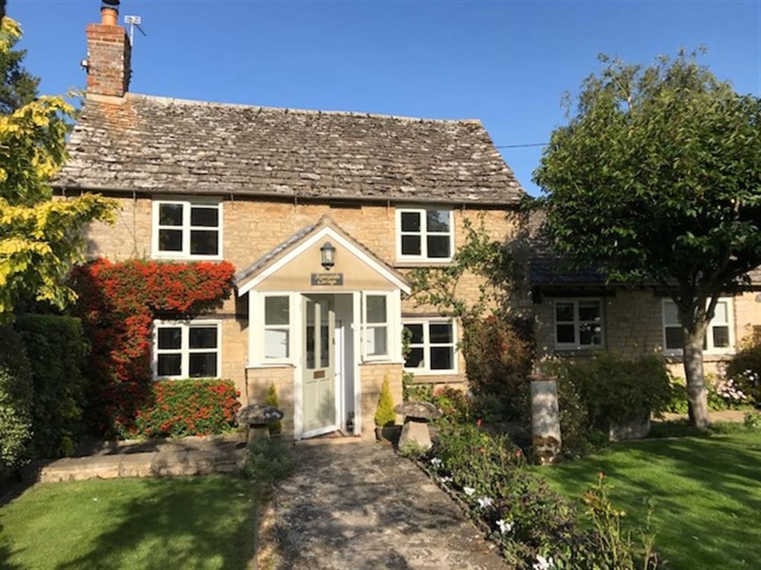 A stone cottage with a chimney a pathway leading to a white door surrounded by plants and trees at Sunnyside Cottage in Bampton