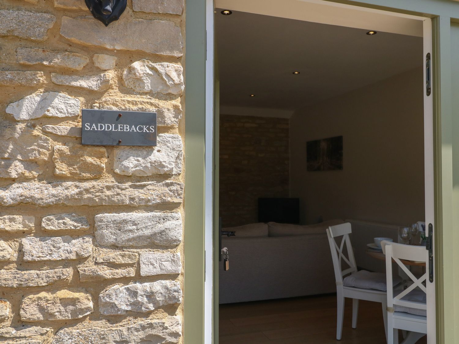 A stone wall with a sign reading saddlebacks and an open door showing a sofa dining table and chairs inside at Saddlebacks Barn Burford