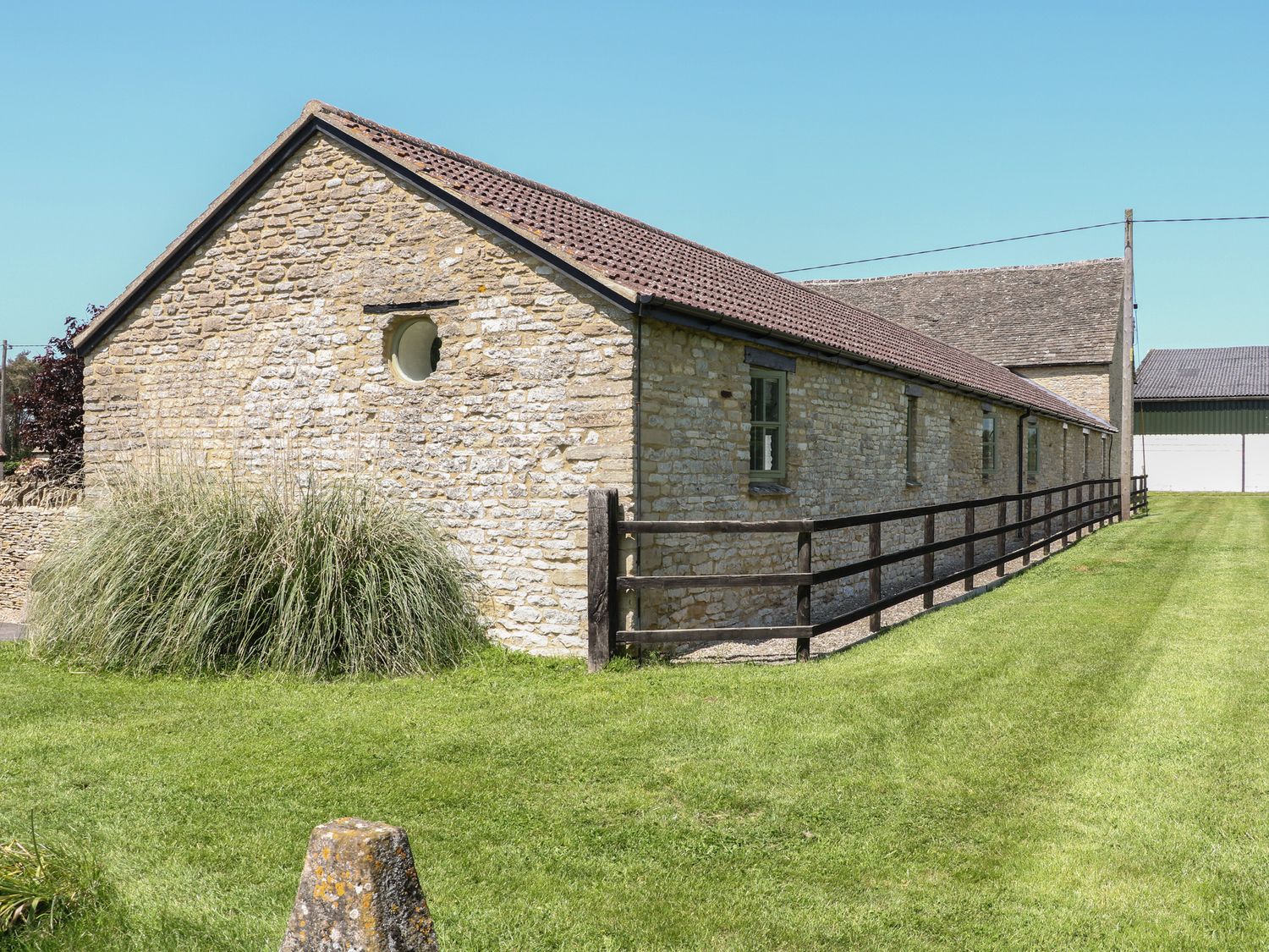 An exterior view of a stone barn with a tiled roof and wooden fence on a grassy lawn at Saddlebacks Barn in Burford
