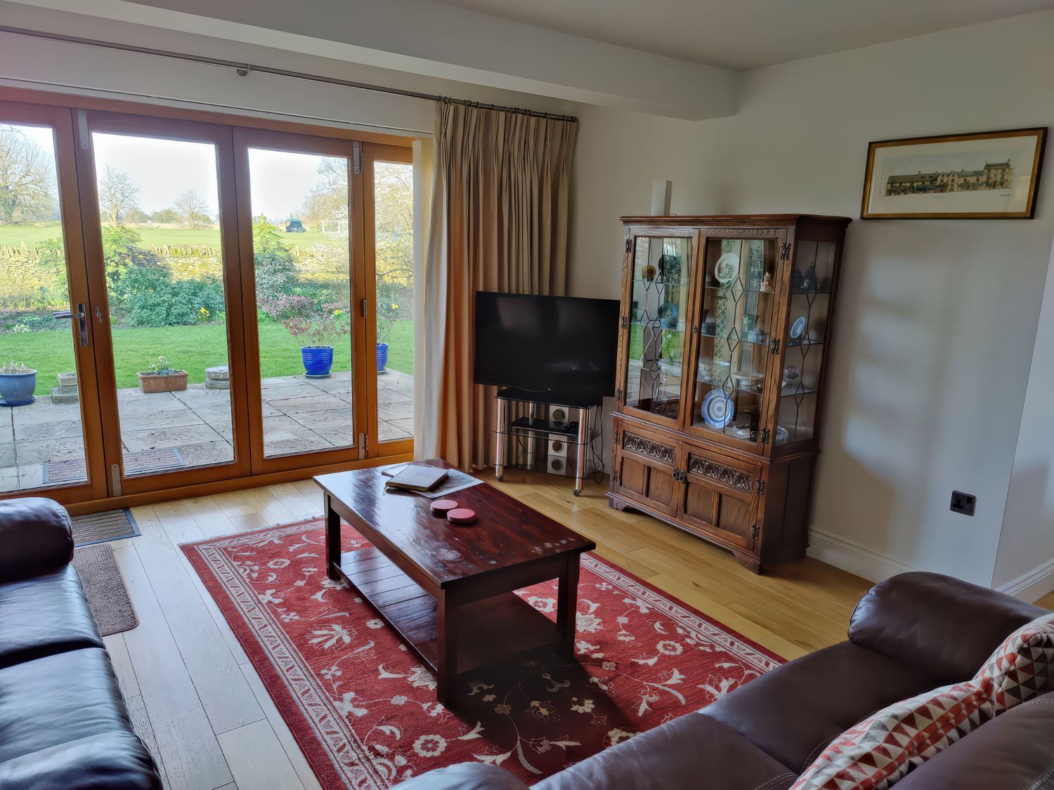 A living room with a wooden coffee table and glass cabinet next to large sliding doors opening to a garden at 5 Burford Mews in Burford