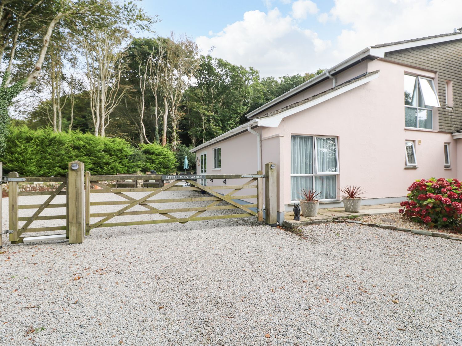 A gravel driveway with a wooden gate and a pink house surrounded by trees at Little Westwards in Goldsithney