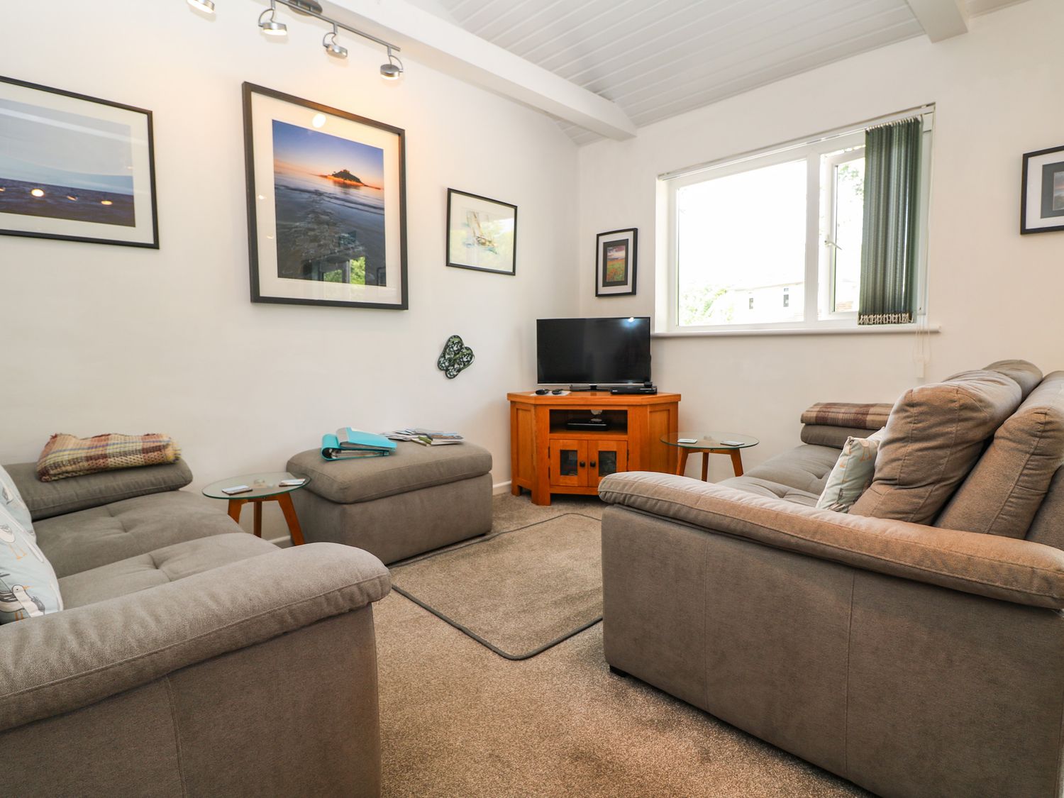 A living room with gray sofas a wooden TV stand with a television glass side tables and framed pictures on the wall at Little Westwards in Goldsithney