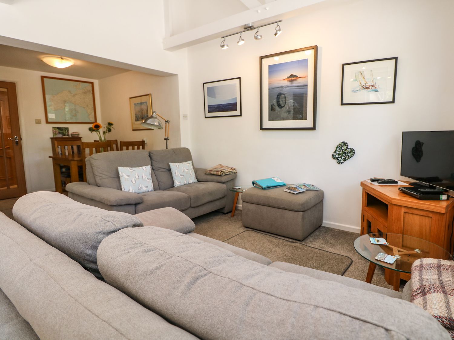 A living room with gray sofas a glass side table a wooden TV stand with a television and framed pictures on the wall at Little Westwards in Goldsithney
