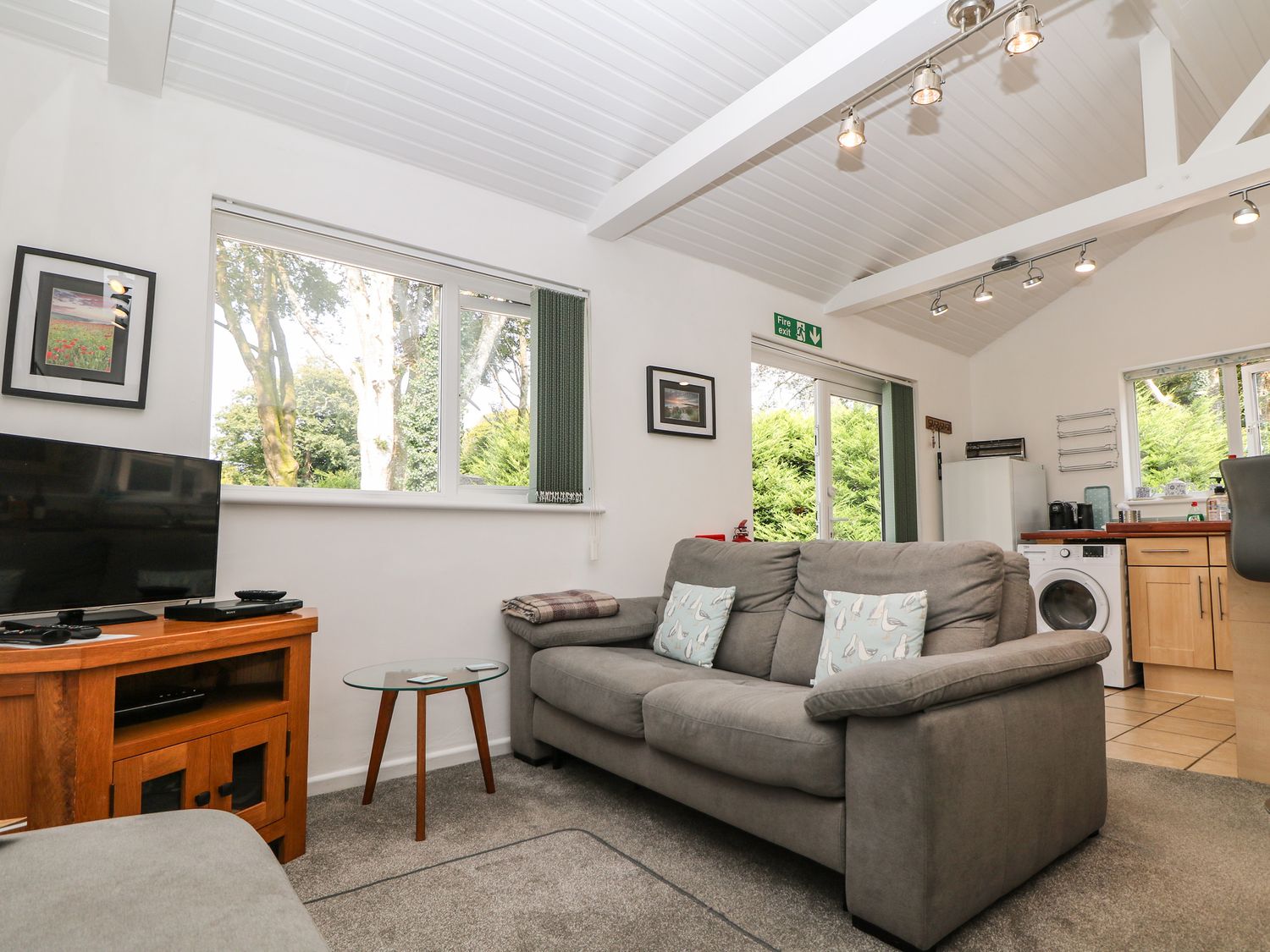 A living room with a gray sofa two cushions a wooden TV stand with a television a glass round side table and kitchen appliances at Little Westwards in Goldsithney