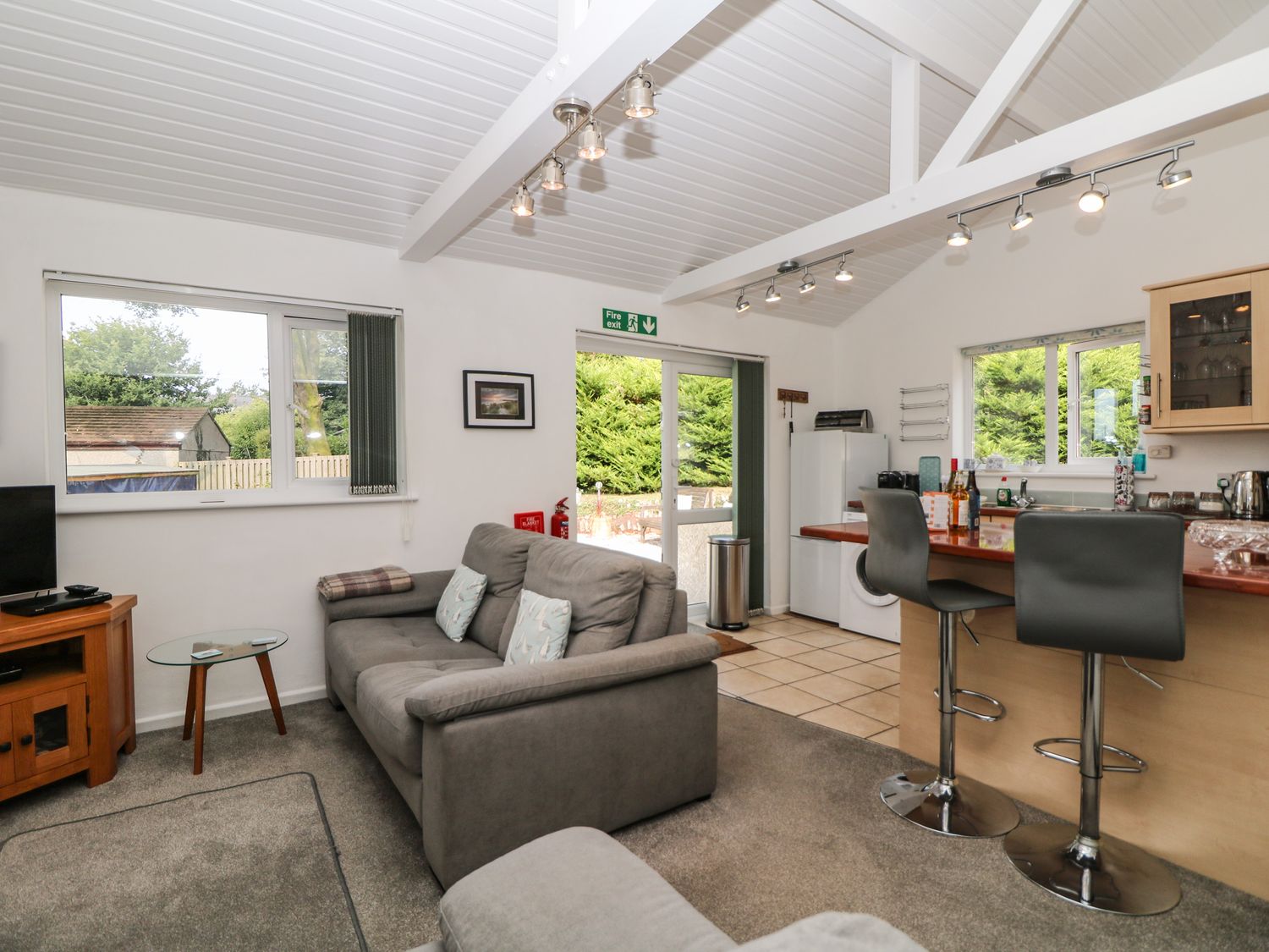 An open plan living area with a gray sofa a small glass side table a tv on a wooden stand a kitchen with a breakfast bar and two stools at Little Westwards in Goldsithney