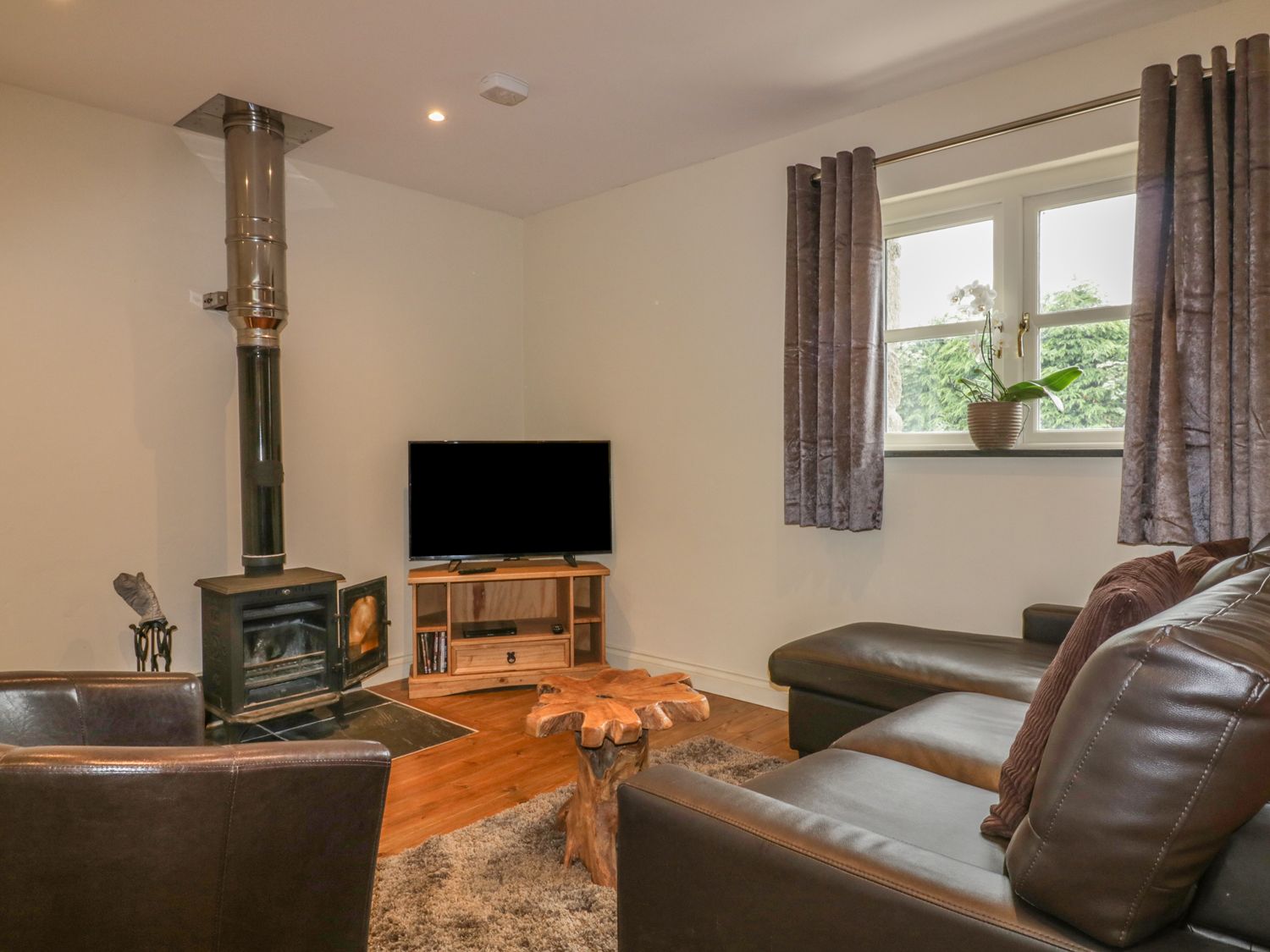 A living room with a wood stove television on a stand leather sofas wooden table and window with curtains at The Smithy in Blisland