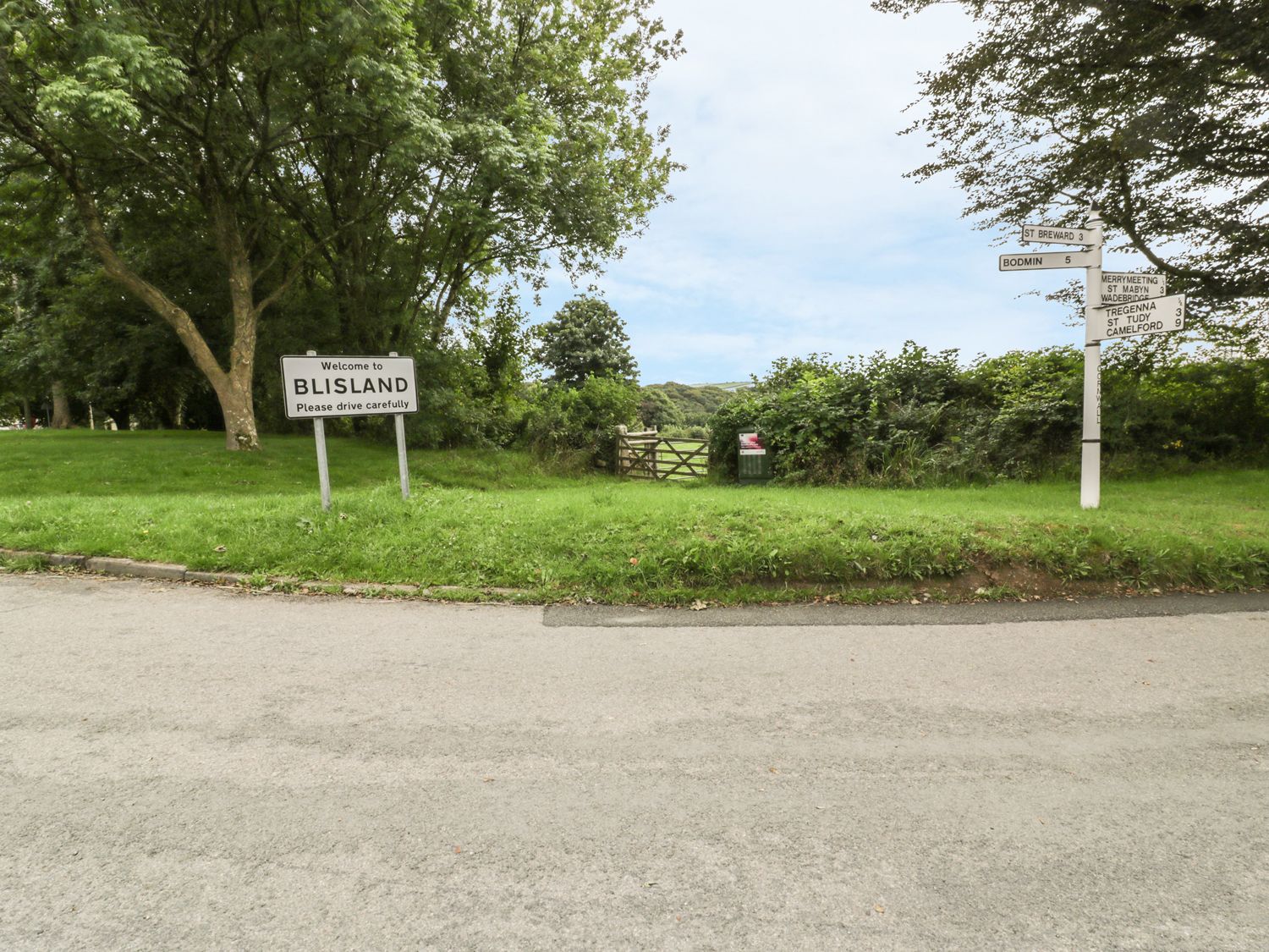 A road with a welcome to Blisland sign and a white signpost next to a grass area with trees at The Smithy in Blisland