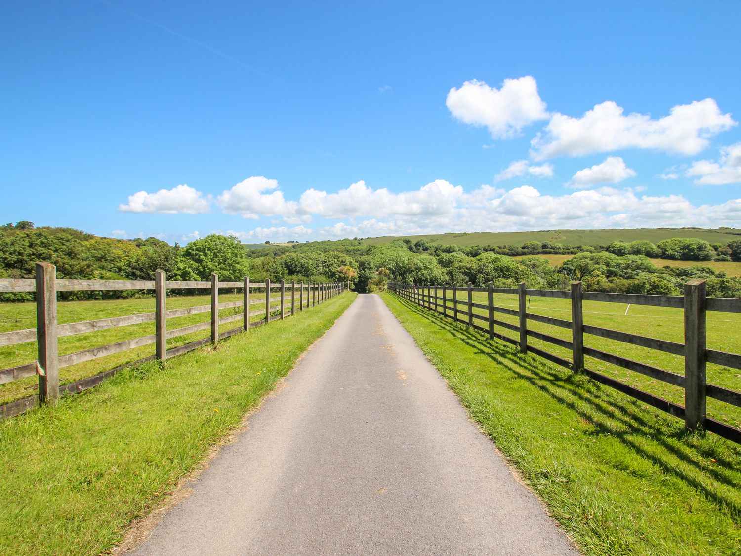A paved road between wooden fences with grass fields and trees under a blue sky at Sunbury Barn in St Agnes