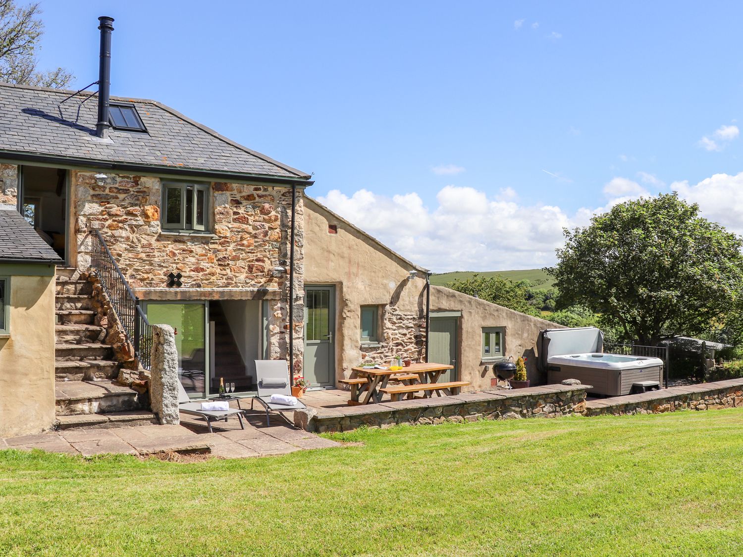 An outdoor patio with lounge chairs a picnic table a barbecue hot tub and grassy lawn at Sunbury Barn in St Agnes