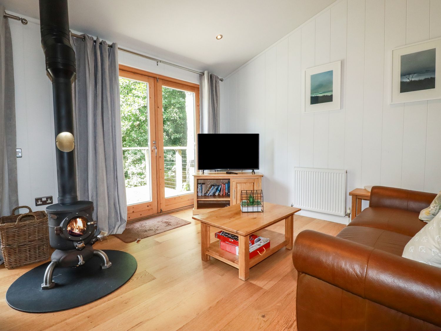 A living room with a wood-burning stove, wooden coffee table, TV on a wooden stand, brown leather sofa, and glass doors at 14 Stonerush Valley Lanreath