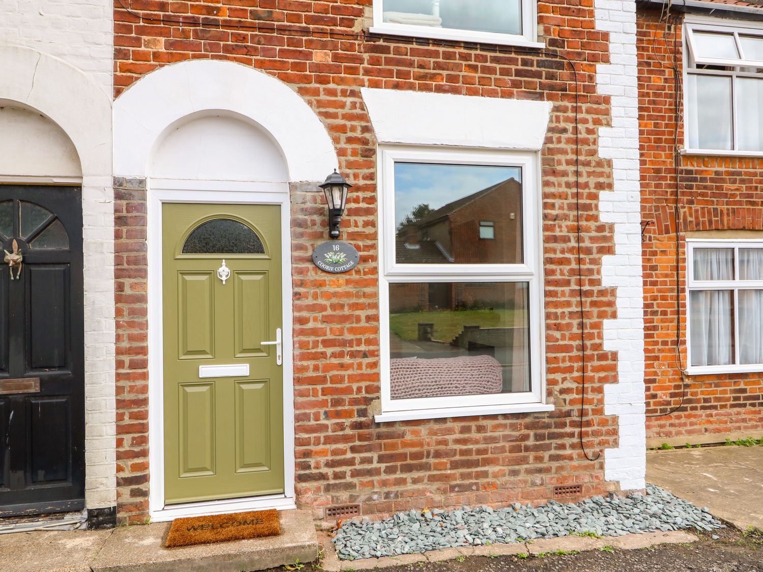 Front door and window of a brick house with a welcome mat and address plaque at Coorie Cottage in Martham