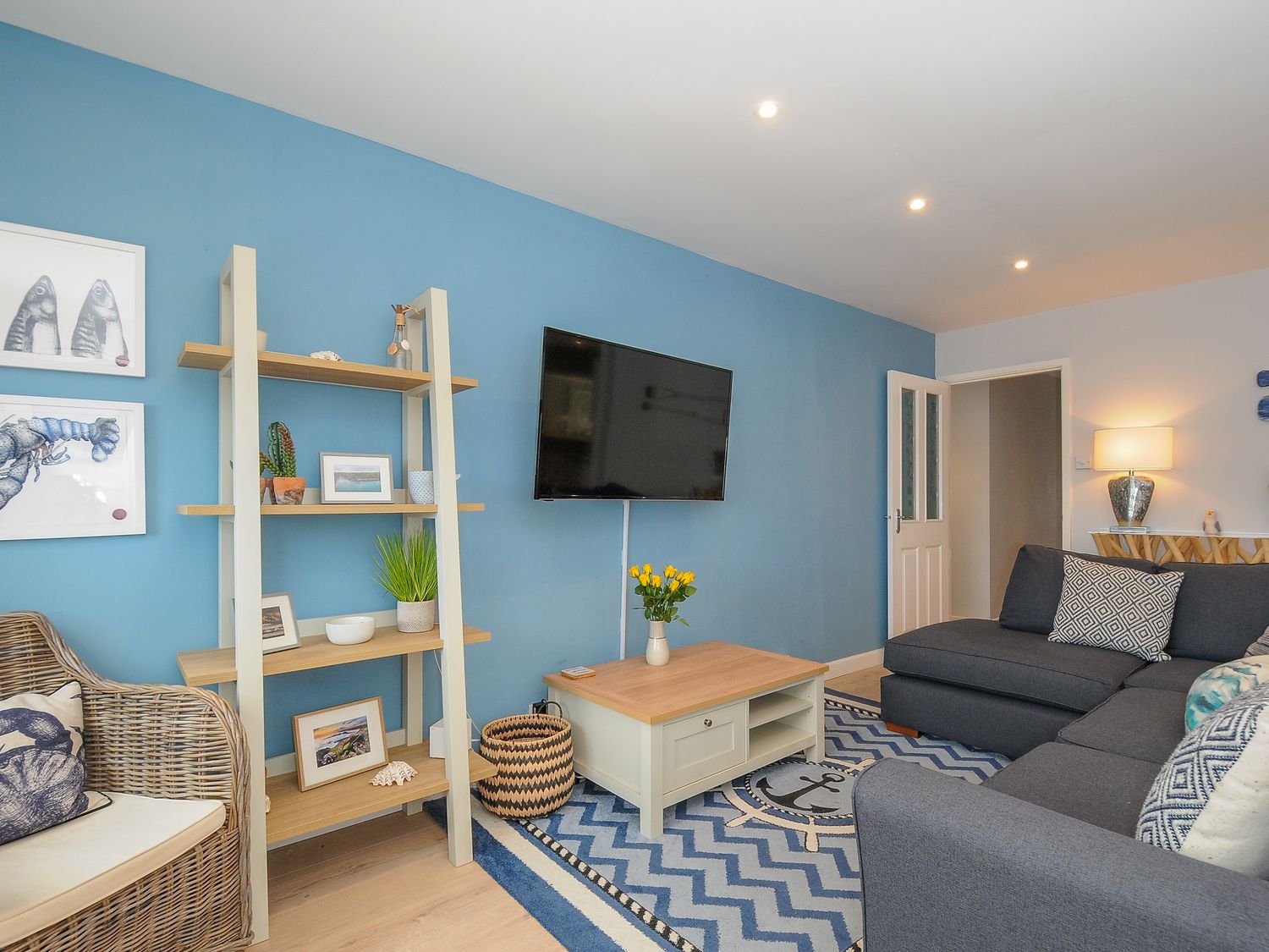 A living room with a blue wall a wall-mounted television a wooden shelf a coffee table and a dark grey sofa at Coastal Haven in Crackington near Crackington Haven