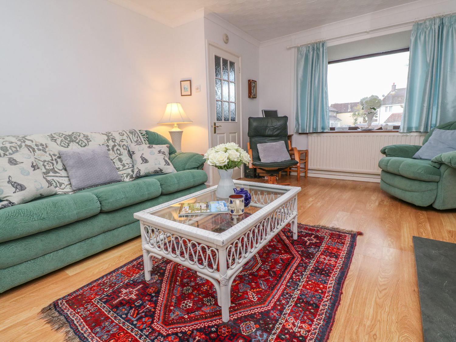A living room with green sofas a wicker coffee table on a red patterned rug and a window with blue curtains at Phenests in Falmouth