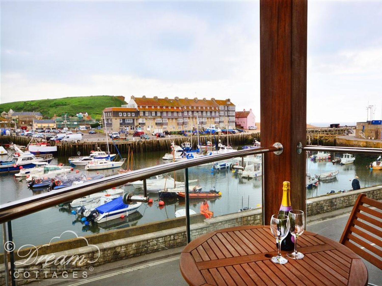 A balcony with a wooden table holding a bottle and two wine glasses overlooking a harbor with boats at The Boardwalk in West Bay