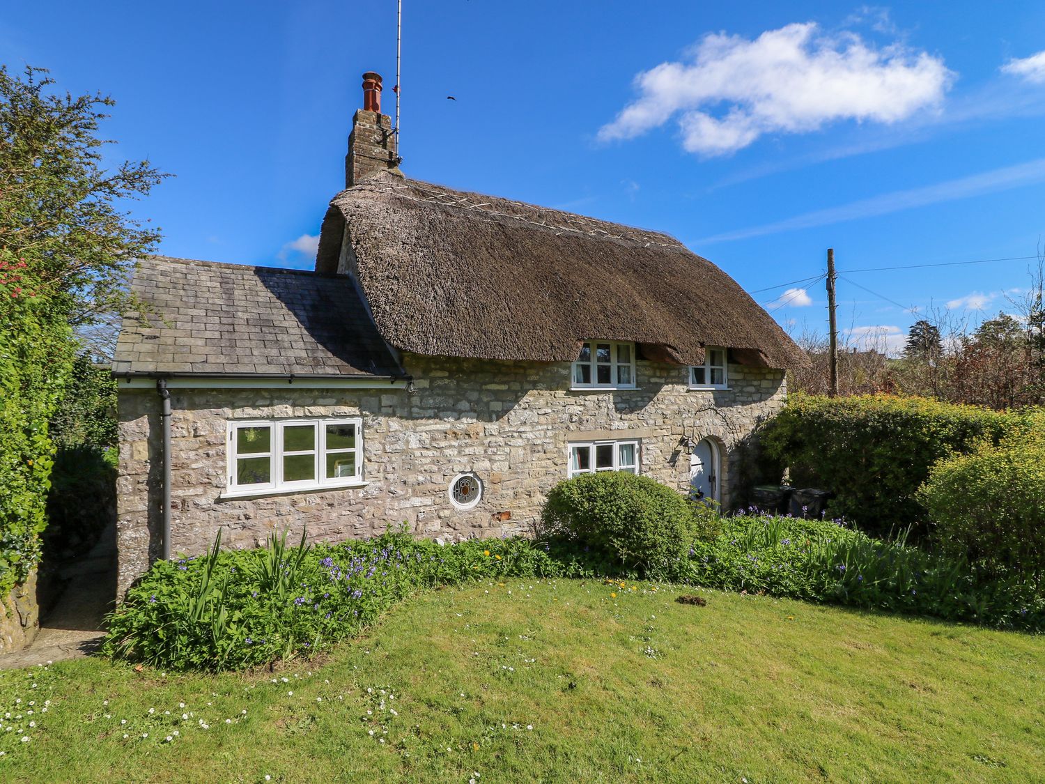 A stone cottage with a thatched roof and garden with bushes and flowers at Lychgate Cottage in Osmington