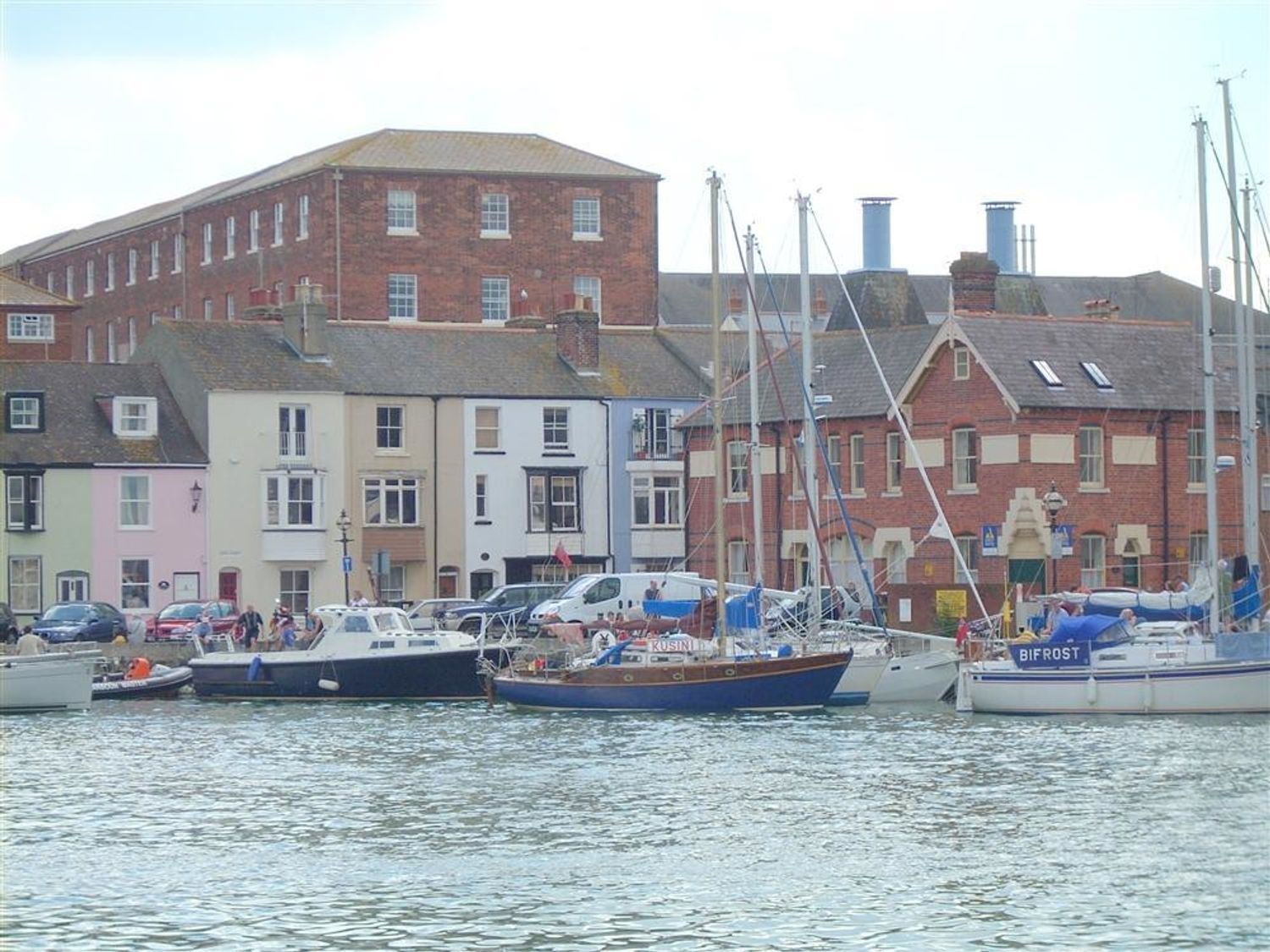 A harbor with several docked sailboats and motorboats by a row of multi-story buildings including brick and pastel-colored houses at Mutiny House in Brewers Quay Harbour