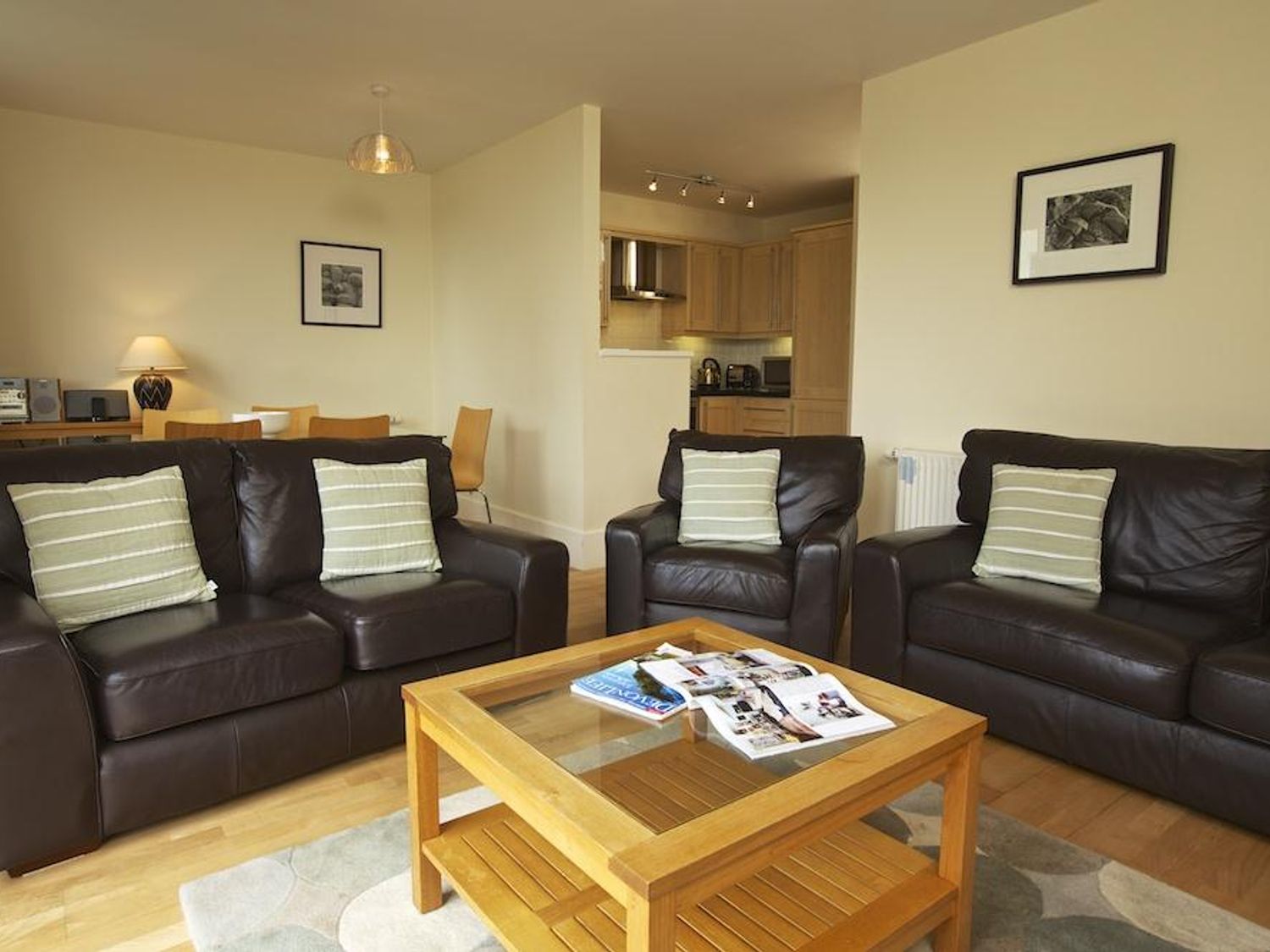 A living room with three dark leather sofas green and white striped pillows a wooden coffee table with magazines and an open plan kitchen at 3 Combehaven in Salcombe