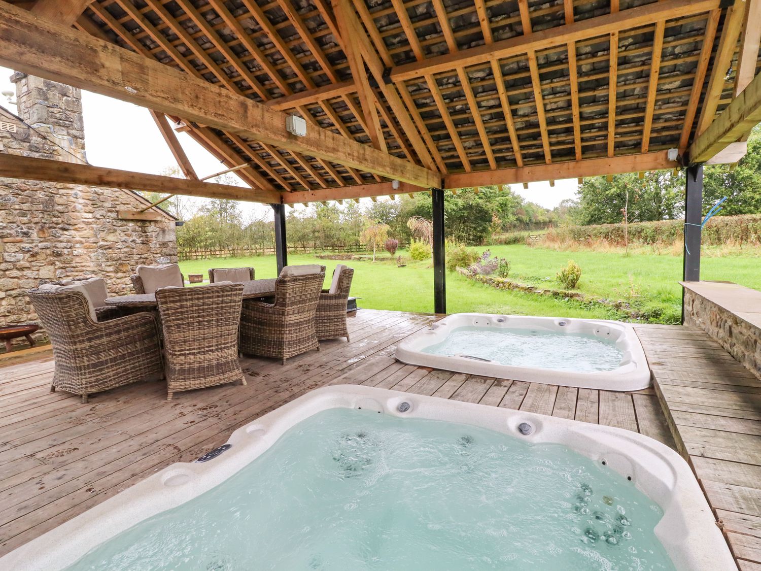 A covered outdoor area with two hot tubs a dining table and wicker chairs overlooking a grass field at Roe Deer Country Retreat in Clitheroe
