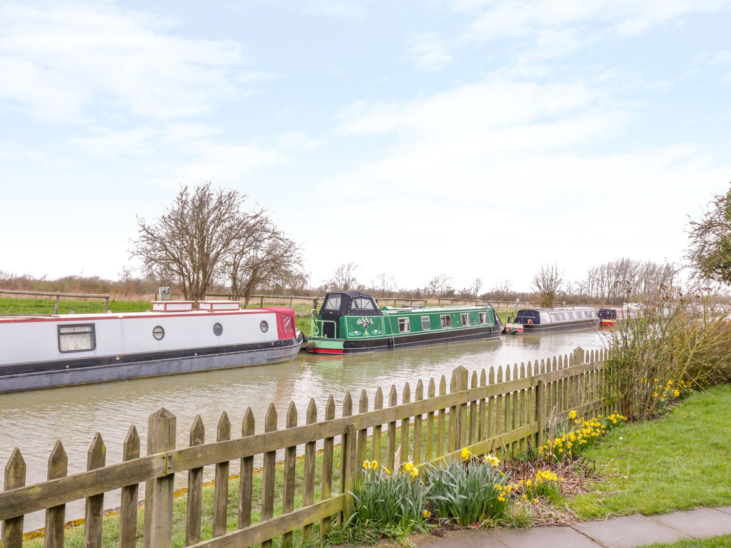 A canal with multiple narrowboats moored near a wooden fence with yellow flowers at Wigrams Canalside Cottage in Napton-on-the-Hill