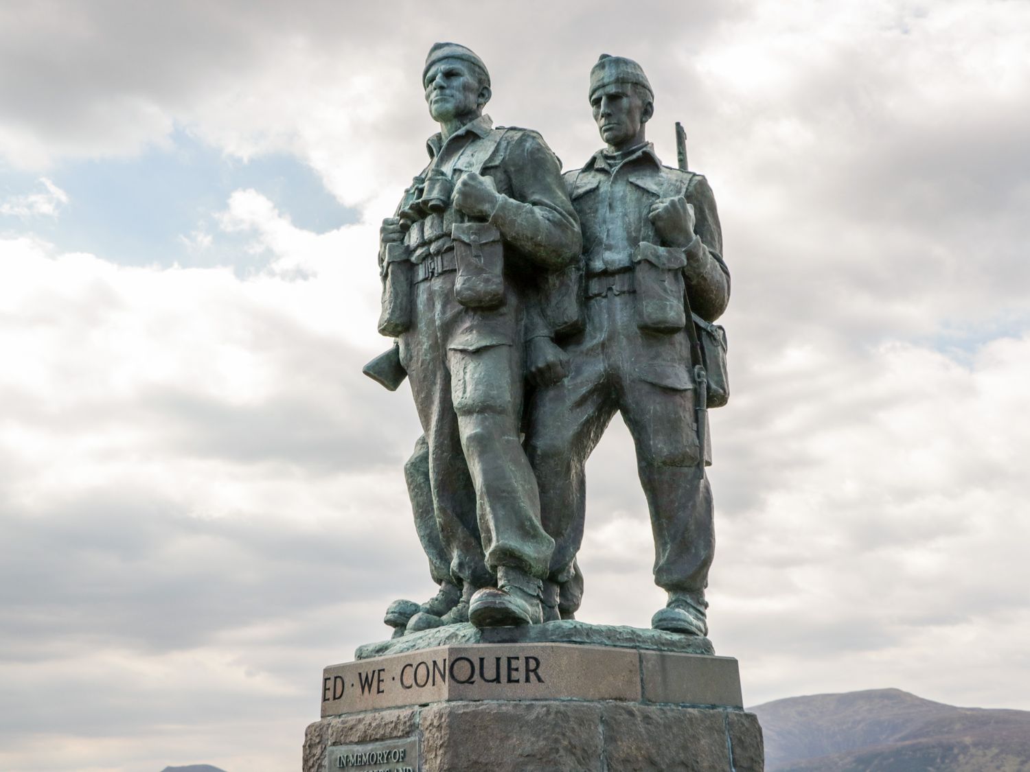 A statue of two soldiers holding weapons on a stone pedestal with mountains and a cloudy sky in the background