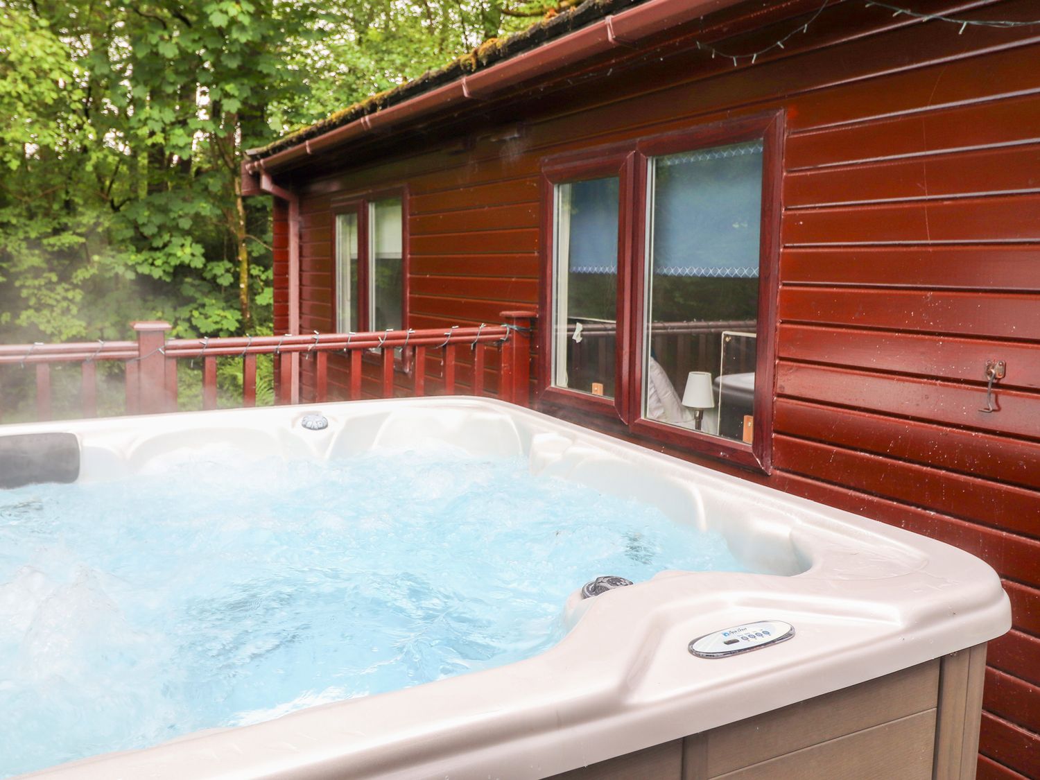 An outdoor hot tub on a wooden deck next to a cabin with red wooden siding and windows overlooking green trees at 40 Skiptory Howe in Troutbeck Bridge
