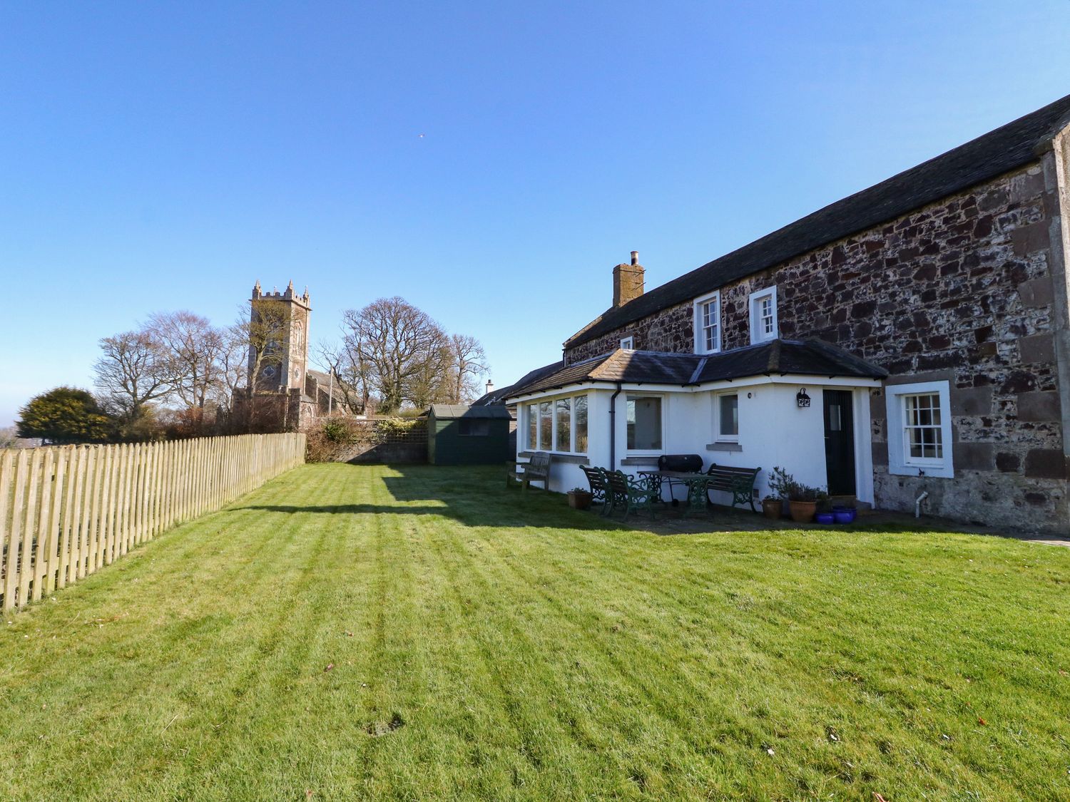 A stone house with a grass yard and a wooden fence near a church at The Old Schoolhouse in Kirkton of Craig near Montrose