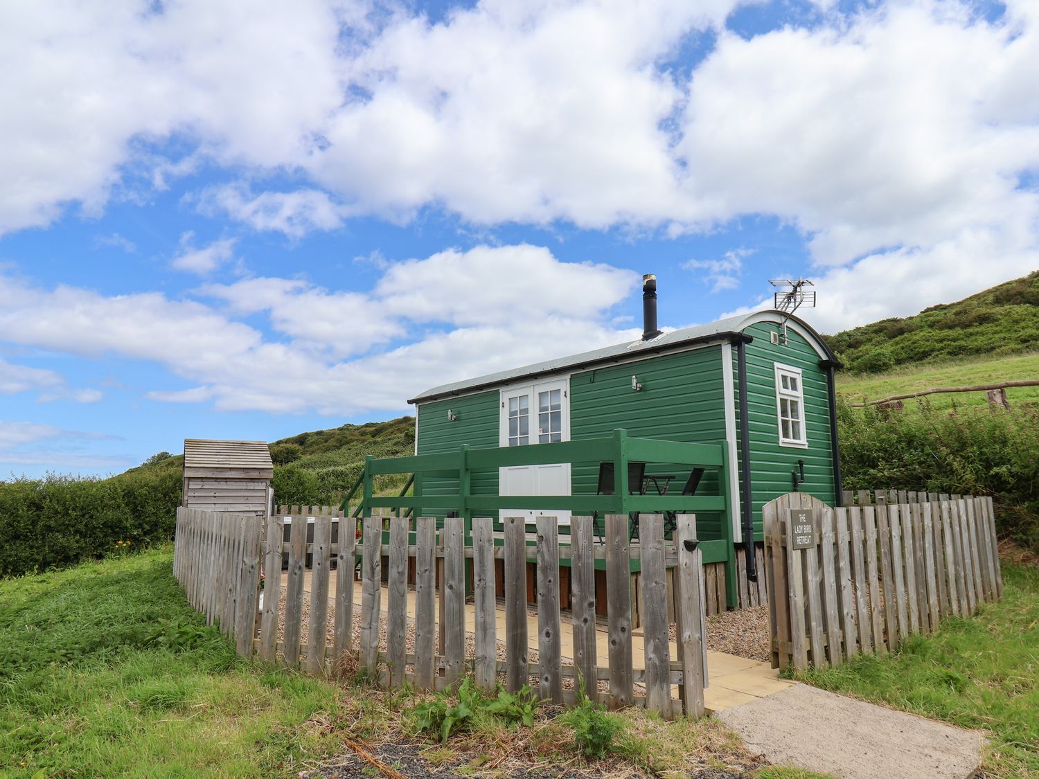 A small green wooden cabin with a fenced yard and chairs on a patio under a partly cloudy sky at Lady Bird Retreat in Skinningrove