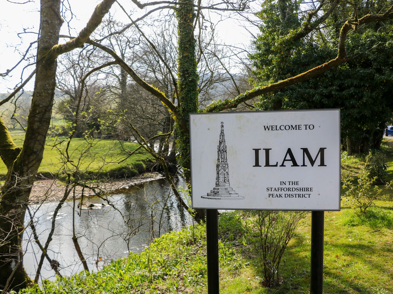 A welcome sign for Ilam near a river with trees and greenery in the Staffordshire Peak District