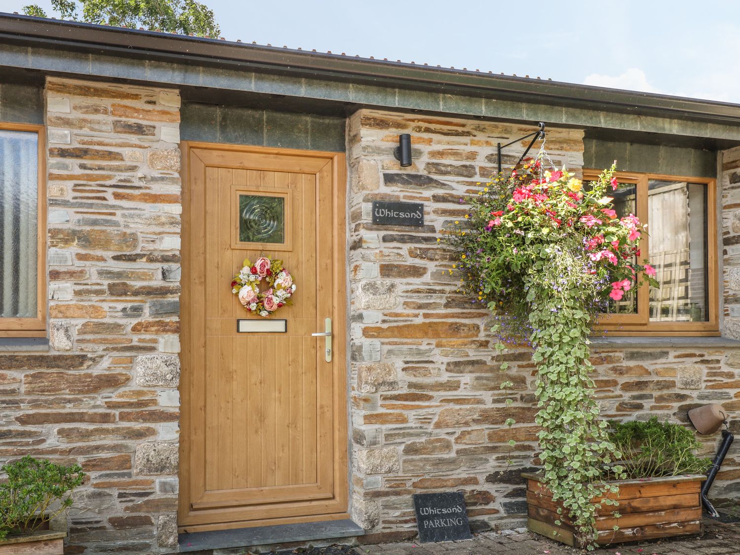 A stone building exterior with a wooden door decorated with a flower wreath and a hanging flower basket at Whitsand in Tideford