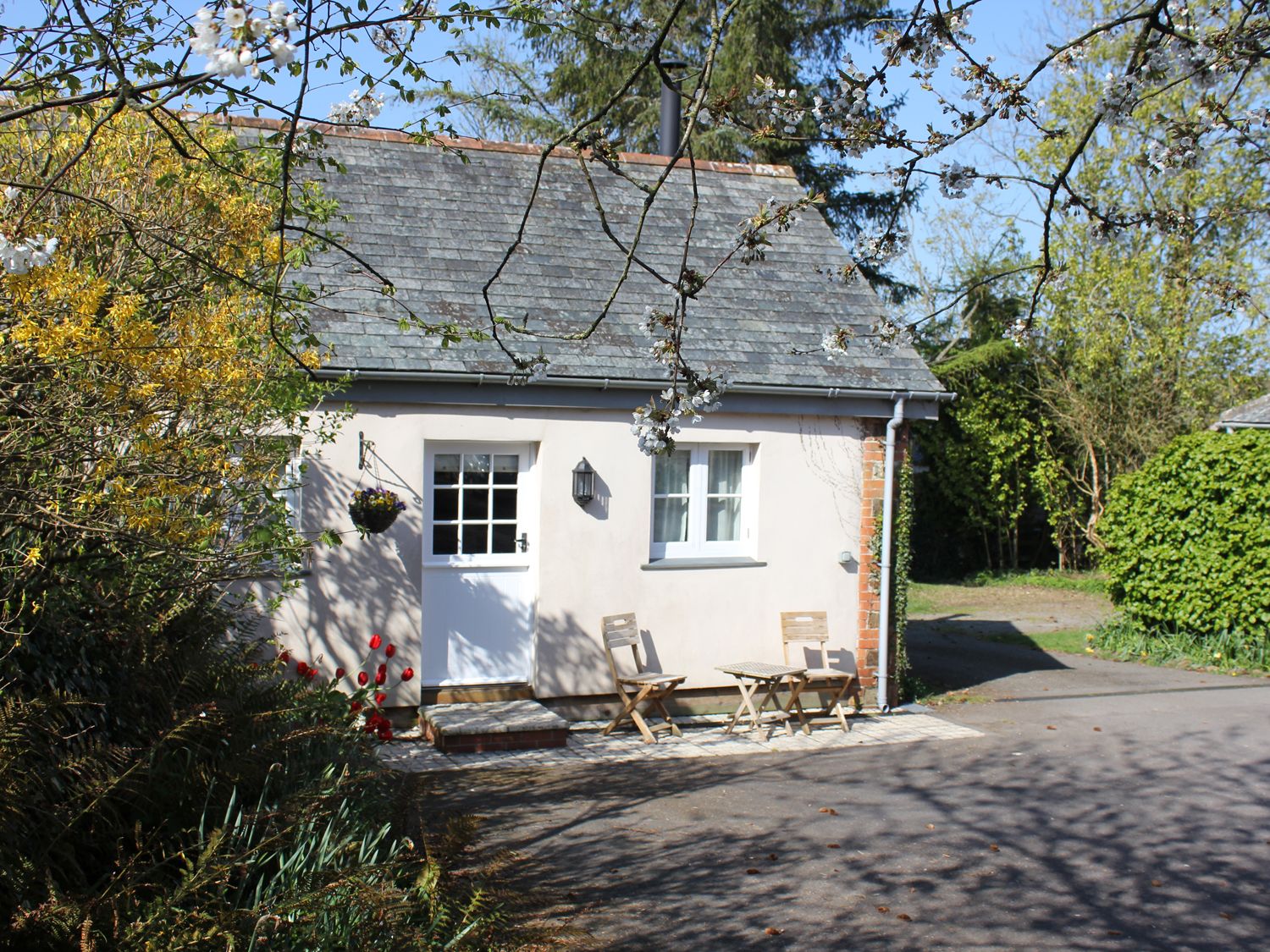 A small house with a white door and window two wooden chairs and a table outside surrounded by trees at The Linney in Littleham