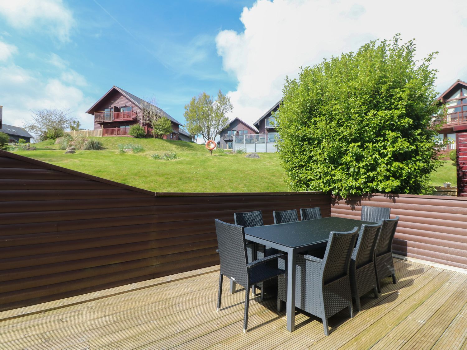 An outdoor deck with a black dining table and six chairs overlooking grassy hills with houses at Lodge Sixty Six in St Columb Major