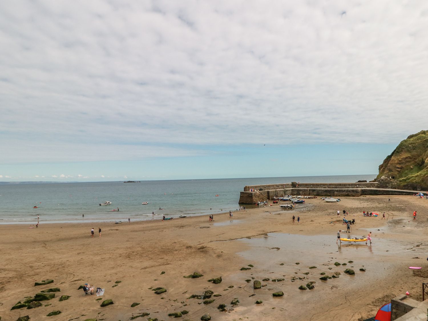 A sandy beach with people walking and boats near a stone pier at Garden House in Gorran Haven
