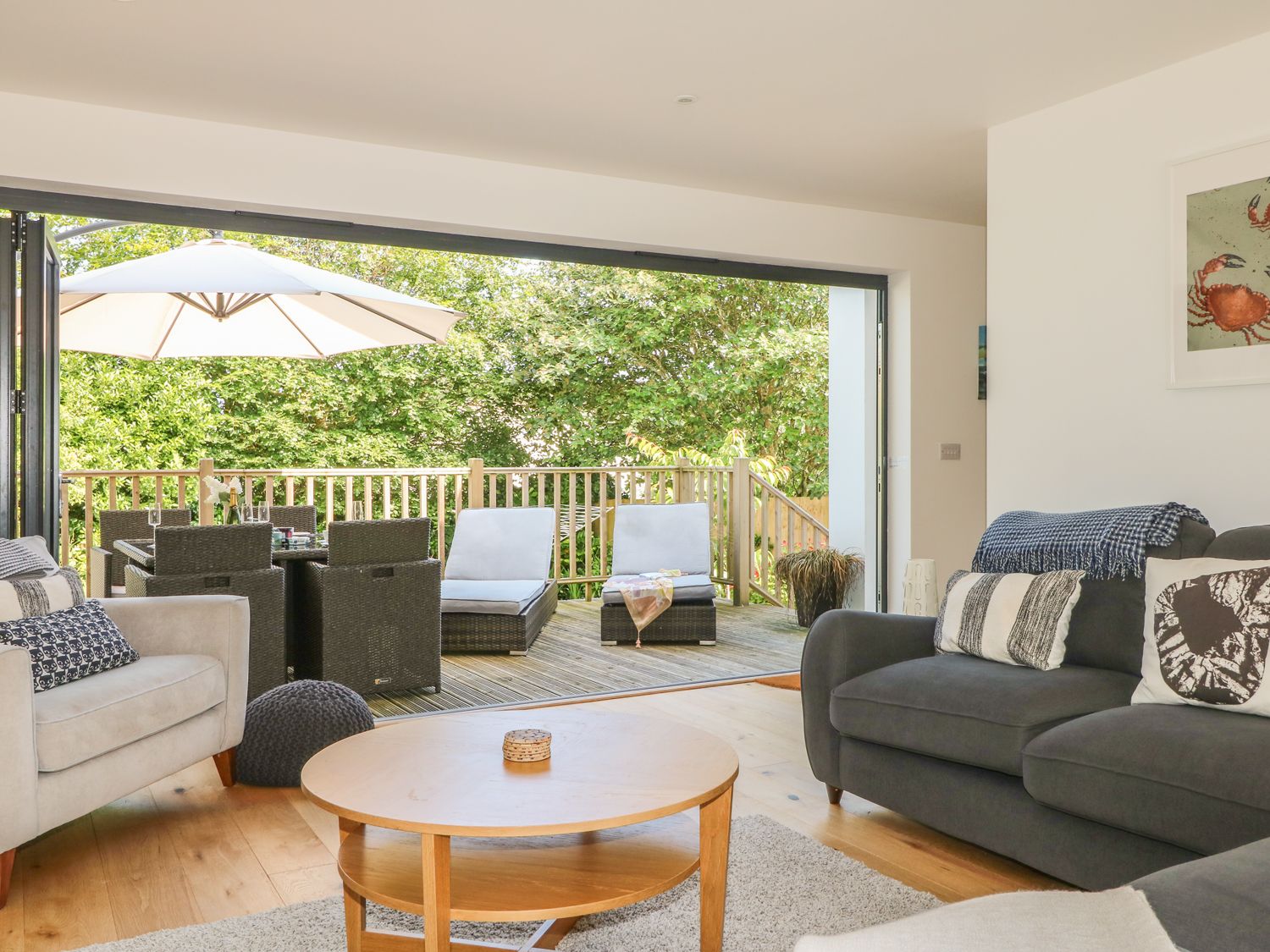 A living room with sofas and a coffee table opening to an outdoor deck with chairs and an umbrella at Garden House in Gorran Haven