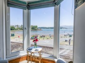 A bay window with a small round table holding a vase of flowers overlooking a waterfront with benches and houses at Bay View Apartment in Porthmadog