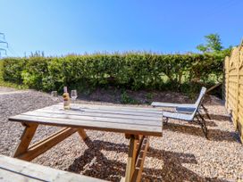 An outdoor area with a wooden picnic table holding a bottle and two glasses and two lounge chairs on gravel near a hedge and wooden fence at Bwthyn Taid in Holywell