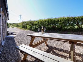 A wooden picnic table with a bottle and two wine glasses on a gravel patio outside a stone building with a hedge and power lines in the background at Bwthyn Taid in Holywell