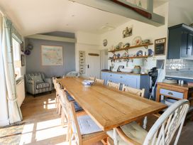 A dining room with a long wooden table surrounded by chairs a striped armchair curtains on windows shelves with pottery and a cabinet at Borth Arian in Rhoscolyn